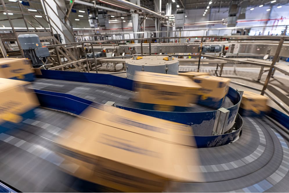 The choice of shipping service providers plays a central role in multichannel e-commerce: belt conveyors in an industrial manufacturing hall with moving cardboard boxes.