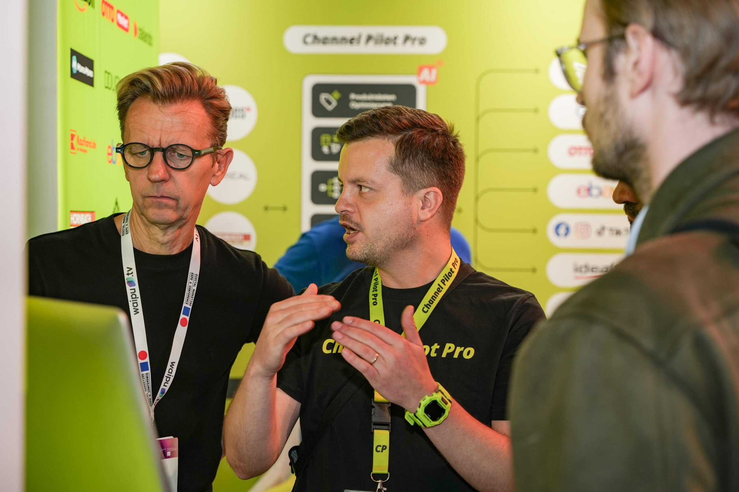 Drei Männer im Gespräch bei einer Fachmesse, hinter ihnen ein grüner Hintergrund mit Logos.